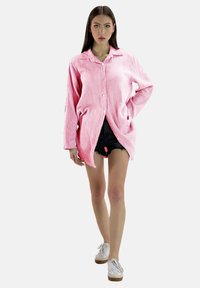 Roze overhemd met een gestructureerd, gestreept patroon, gecombineerd met zwarte denim shorts. Model draagt witte sneakers.