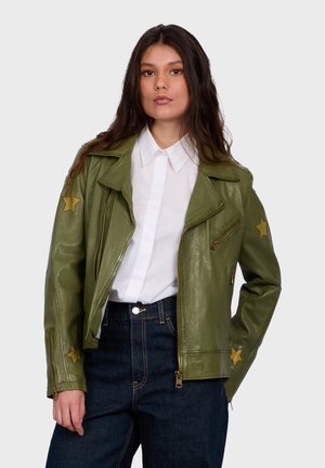 Woman wearing an olive green leather jacket with yellow star patches on sleeves, white button-up shirt, and dark blue high-waist jeans.