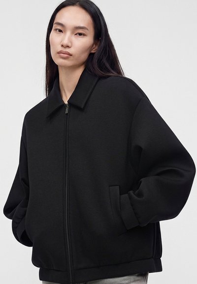 Femme aux longs cheveux foncés portant une veste noire ample à fermeture éclair, les mains dans les poches, sur un fond clair uni.