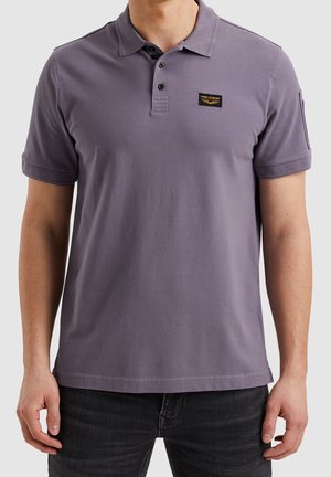 Man draagt een paars poloshirt met korte mouwen, zwarte knopen en een klein PME Legend-logo op de borst, gecombineerd met een zwarte spijkerbroek.