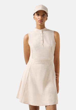 Kobieta ubrana w beżową sukienkę bez rękawów z guzikami z przodu i dopasowaną beżową czapkę z daszkiem, stojąca na prostym jasnym tle.