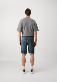 Cămașă gri cu mânecă scurtă asociată cu pantaloni scurți din denim albastru, cu manșete răsucite, și adidași albi, văzută din spate pe un fundal simplu.