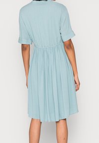 Ljusblå klänning med korta uppvikt ärmar, midjeband och vertikala veck, tillverkad av ett mjukt, lätt material.