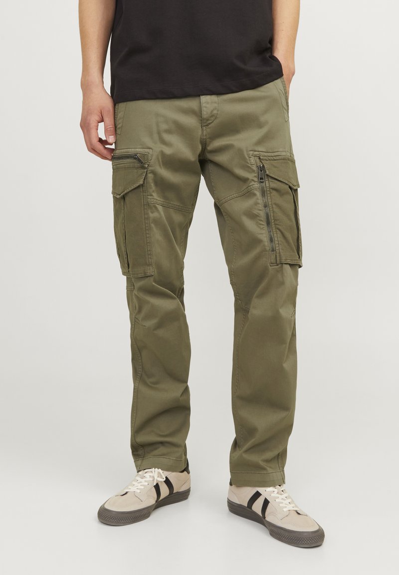 Cotton cargo trousers in olive green with multiple pockets, zippered details, and a loose fit, paired with beige sneakers featuring black stripes.
