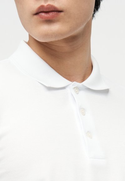 Κοντινό πλάνο στο κάτω μέρος του προσώπου και τον λαιμό ενός ατόμου που φοράει λευκό πόλο πουκάμισο με τρία κουμπιά κλειστά, μπροστά σε απλό φόντο.
