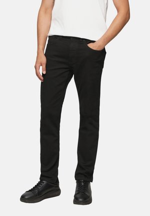 Mężczyzna w czarnych dopasowanych jeansach z ręką w kieszeni i czarnych tenisówkach, stojący na jednolitym tle.