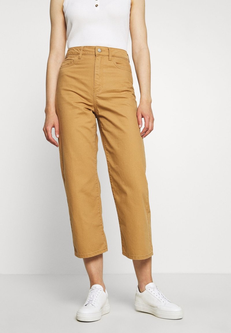 Jean cropped beige en coton, taille haute, avec fermeture à bouton et poches avant. Porté avec des baskets blanches. Fond uni.