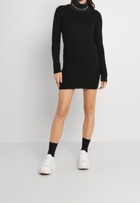 Abito lungo nero a maglia con un motivo testurizzato, silhouette aderente e polsini a costine. Abbinato a sneakers bianche e calzini neri.