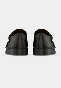 Sapatos de pele preta com um acabamento suave, apresentando um perfil baixo e dois fechos de fivela nas laterais. Sola plana de borracha.