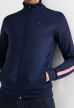 Mand iført marineblå lynlåstrøje med Tommy Jeans-logo på brystet og mærket stribe langs ærmerne, hænderne i lommerne.