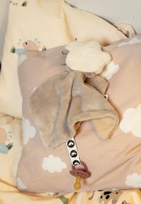 Suave mantita de peluche beige con cabeza blanca esponjosa de corderito, chupete adherido en una etiqueta de tela, colocada sobre una almohada marrón claro con patrón de nubes.