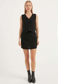 Svart texturerad väst med tre framtill knappar och matchande figursydd kjol. Modell bär svarta ankelboots som framhäver en kort design.