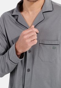Grijze pyjama-top met een notch-kraag, zwarte piping, voorzak en knopenluiting. Gemaakt van zacht materiaal met een gladde textuur.
