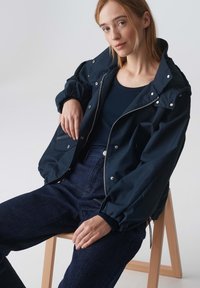 Marineblauwe bomberjack met een rits aan de voorkant, drukknoopsluiting en elastische manchetten. Draag het over een zwart geribbeld topje, gecombineerd met donkere spijkerbroek.