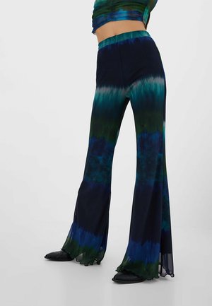 Frau trägt hoch taillierte, ausgestellte Hose mit blau-grün-schwarzem Batikmuster und schwarzen Schuhen vor schlichter Hintergrund.