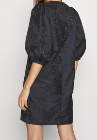 Černé šaty se saténovou texturou, vycpanými rukávy a složitým tmavým vzorem. Mají šev na zadní straně a uvolněný, áčkový střih.