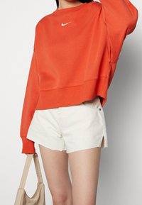 Pomarańczowa bluza z prążkowanymi mankietami i dołem, z małym białym logo Nike. W połączeniu z białymi szortami jeansowymi, odsłaniającymi boczne rozcięcia.