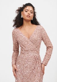 Sukienka z długim rękawem, ozdobiona cyrkoniami, w delikatnym różu, z dekoltem w kształcie V, posiadająca fason kopertowy i marszczenia w talii.