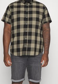 Camisa de manga corta de cuadros en verde oliva y negro, con cuello abotonado y dobladillo redondeado, combinada con pantalones cortos de mezclilla gris.