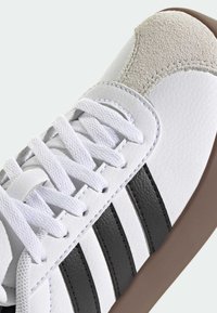 Biały sneaker z czarnymi paskami, białymi sznurowadłami, szarą zamszową osłoną palców i brązową gumową podeszwą, ustawiony pod kątem, aby pokazać szczegóły boczne i górne.