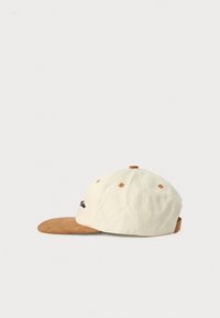 Beige six-panel cap with brown suede brim, brown button and eyelets, and small black embroidered text on the front left side.