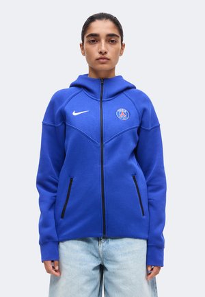 Femme portant un sweat à capuche zippé bleu Nike avec le logo Paris Saint-Germain, associé à un jean bleu clair, debout devant un fond uni.