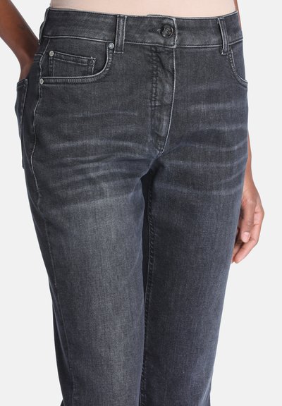 Gros plan d'une personne portant un jean denim gris foncé avec des poches avant et une fermeture par bouton, main reposant sur le côté.