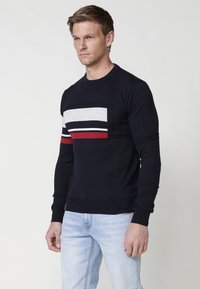 Pulover tricotat navy cu un design geometric, având dungi orizontale gri, alb și roșu pe piept și pe mâneci. Manșete ajustate.