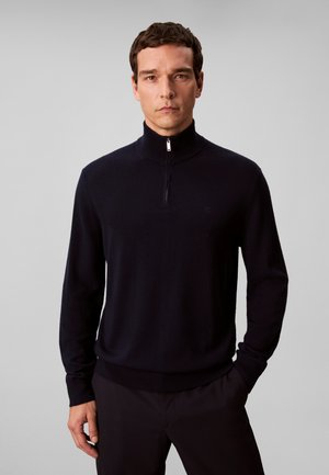Man die een zwarte trui met lange mouwen en halve rits draagt en een zwarte broek, staand tegen een effen lichtgrijze achtergrond.