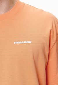 Primer plano de una persona que lleva una camisa de color melocotón con el texto blanco "PEGADOR" impreso en el pecho.