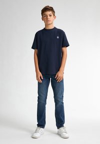 T-shirt en coton bleu marine à manches courtes avec un logo en patch sur la poitrine, associée à un jean bleu ajusté et des baskets blanches sur un fond uni.