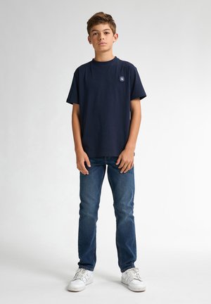 Ragazzo adolescente in piedi, indossa una t-shirt blu navy con un piccolo logo, jeans blu e sneakers bianche su uno sfondo chiaro e uniforme.