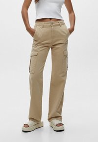 Pantalon cargo beige à coupe décontractée, avec poches latérales, braguette zippée et devant plat. Porté avec des sandales à plateforme de couleur claire.