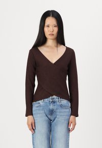 Haut marron à manches longues avec un décolleté en V et un design croisé, présentant un tissu texturé. Porté avec un jean bleu clair à coupe décontractée.