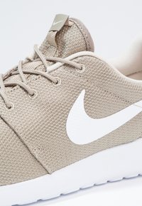 Ljusbeige Nike-sneakers med vitt swoosh-logotyp, meshtopp, beige snören och vit sula visade i närbild från sidan.