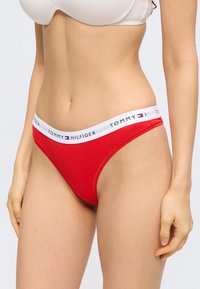 Γυναίκα που φορά κόκκινο εσώρουχο μπικίνι Tommy Hilfiger και μπεζ σουτιέν, εμφανιζόμενη από τη μέση έως τους μηρούς πάνω σε μονόχρωμο φόντο.