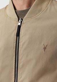 Chaqueta bomber color beige con una textura suave, cuello acanalado, cierre de cremallera y un motivo bordado que presenta un detalle de calavera con cuernos.