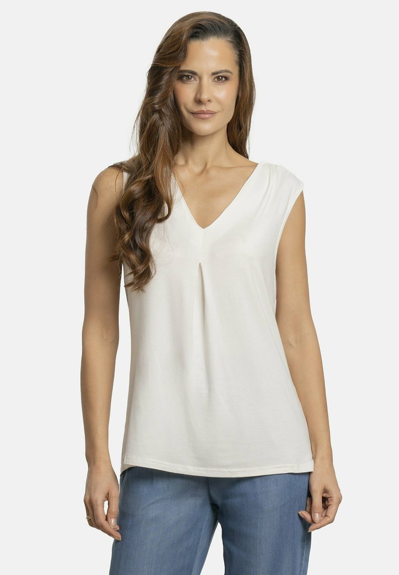 Top sin mangas de cuello en V en tela color crema, con un plisado en el pecho y textura suave. Combinado con pantalones de mezclilla azul claro.