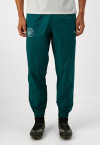 Pantaloni leggeri color turchese con polsini elasticizzati, dotati di tasche laterali e logo del Manchester City sulla coscia sinistra. Tessuto dalla superficie liscia.