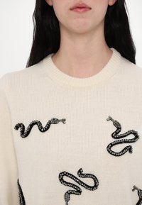 Pūkains off-white džemperis ar melnām čūskām, kuras veidotas ar dekoratīviem rhinestones, apaļa izgriezuma kakla un ribotas apkakles detaļām.