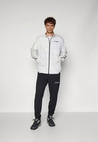 Witte vest met ritssluiting en patroon, met zwarte accenten, gecombineerd met zwarte joggingbroek met logo, aangevuld met zwart schoeisel.