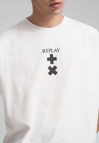 T-shirt in cotone bianco con una stampa grafica nera con la parola "REPLAY" sopra due simboli. Vestibilità ampia con scollatura rotonda.