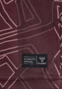 Tessuto marrone con motivi geometrici. Un'etichetta nera riporta la scritta "Authentic Apparel" con un logo a forma di ape, che indica il marchio Hummel in lettere bianche.