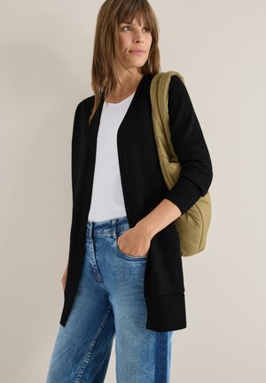 Schwarze, offen gestrickte Strickjacke mit langen Ärmeln, getragen über einem weißen T-Shirt und blauen Jeans. Eine hellolivgrüne Tasche hängt über einer Schulter.