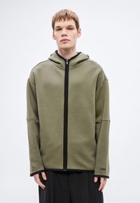 Olivovo zelená mikina s kapucňou na zips, čiernym zipsom, priestrannou kapucňou a dlhými rukávmi s čiernymi manžetami. Logo "BOSS" na zápästí.