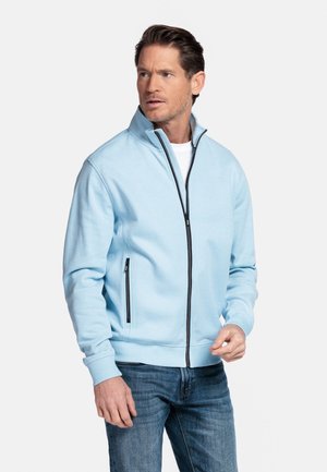 Mann trägt eine hellblaue Jacke mit Reißverschluss über einem weißen Hemd und blauen Jeans, schaut mit neutralem Gesichtsausdruck zur Seite.