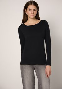 Schwarzes Langarmshirt mit U-Boot-Ausschnitt, aus weichem Stoff gefertigt. Kombiniert mit hellgrauen Jeans steht das Model vor einem neutralen Hintergrund.