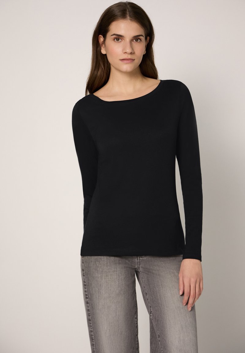 Schwarzes Langarmshirt mit U-Boot-Ausschnitt, aus weichem Stoff gefertigt. Kombiniert mit hellgrauen Jeans steht das Model vor einem neutralen Hintergrund.