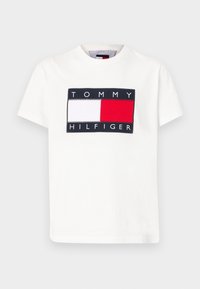 Valkoinen puuvillapaita, jossa on suorakulmainen logomuotoilu, jossa on laivastonsinisiä, punaisia ja valkoisia osia. Brändätty "TOMMY HILFIGER" -tekstillä, joka on kirjoitettu lihavoituna.