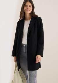 Manteau noir au design ajusté, deux boutons, revers crantés et poches latérales. Porté par-dessus un haut clair avec un jean gris et un sac.